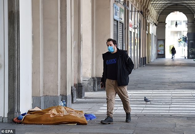 A man wearing a protective mask walks past a homeless asleep in his sleeping bag under one of the arcades in the center of Turin, Italy, today
